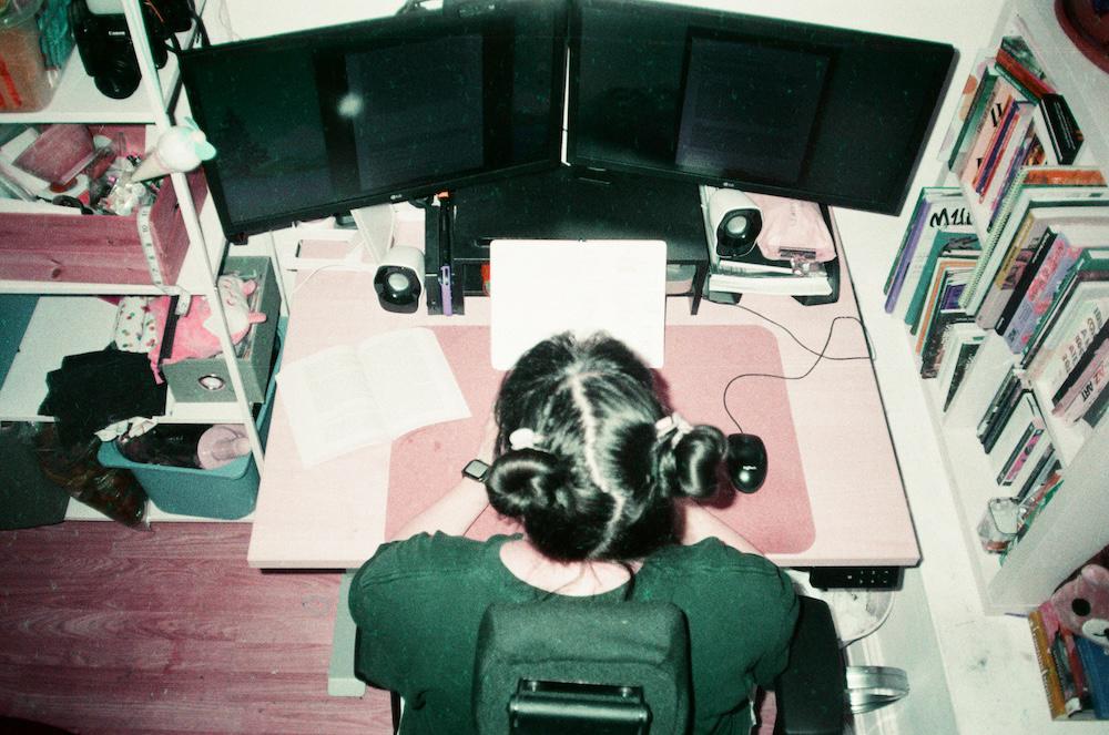 an overhead shot of gab with space buns working at a two screen desk in between shelves of books and sewing materials