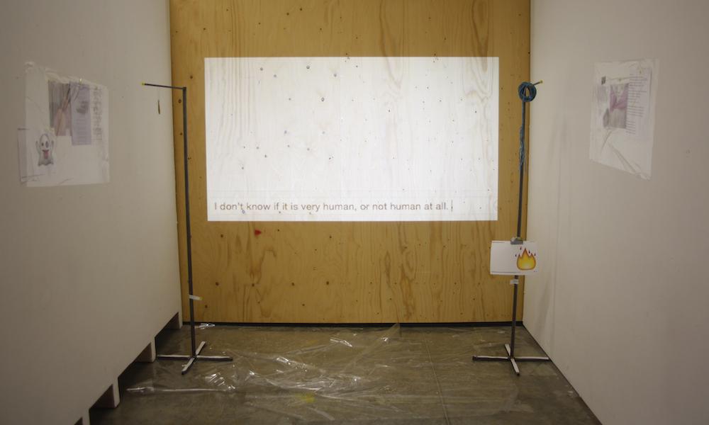 a white projection on the wall says i dont know if it is very human or not human at all, mid video, plastic on the floor below, two metal stands holding objects including wire and a print out of the fire emoji, with further plastic on the walls to the side holding up lyrics and blurred out pictures