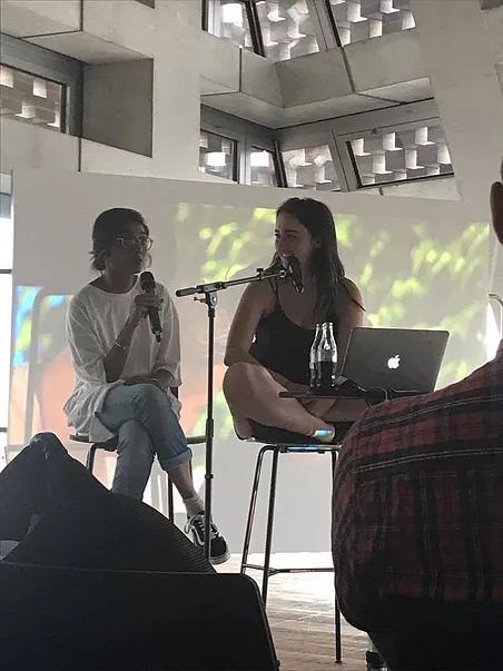 gabrielle sits cross legged with a laptop in front of her while she smiles at zarina on a stool next to her with the mic