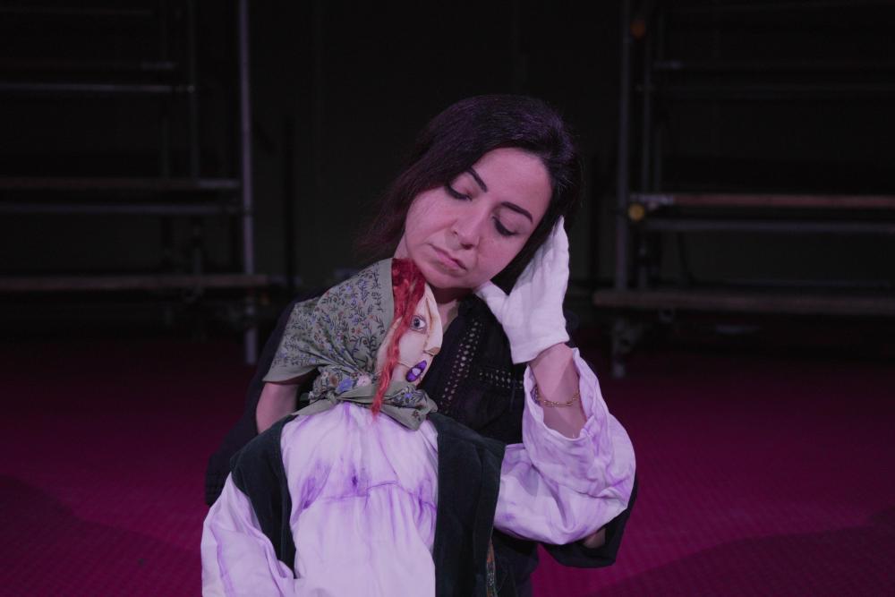 mahtab performing on stage with a puppet in front of her, raising an arm to touch her face