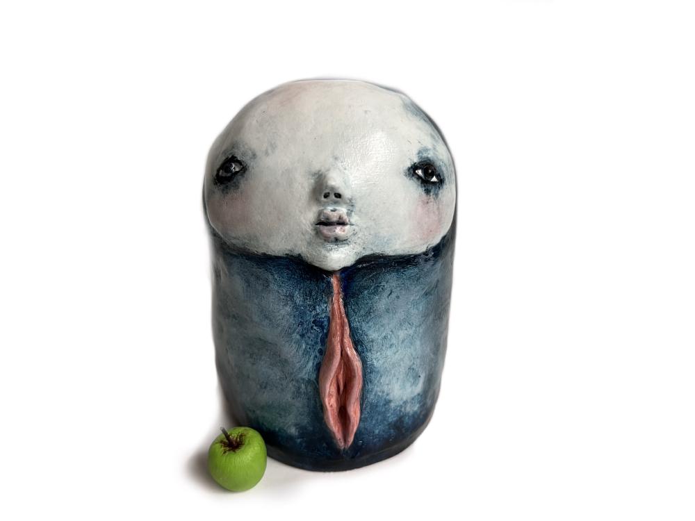 a round sculpture, stubby, with a wide ashen face, a blue body, and labia down its front, next to an apple on a plain background