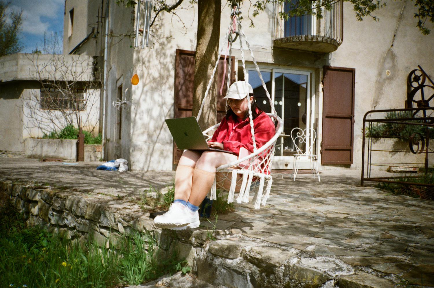 Gab writing on a laptop in a swing attached to a tree with the mill building in the background