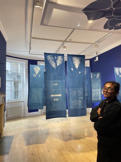 blue cyanotype flags hanging in aa gallery showing ursula k le guin's maps, plus zarina with her arms folded to one side of the picture