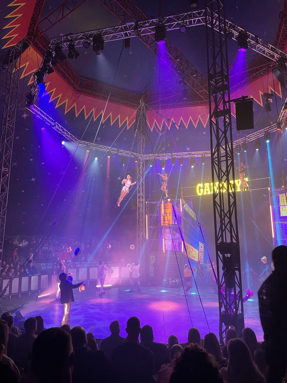 a woman high up and standing on a swing in the circus with men on the ground tossing hats